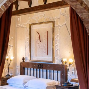 Une chambre avec un mur en pierre, une œuvre d’art encadrée, une tête de lit en bois, deux tables de chevet avec des lampes et un lit avec du linge de lit blanc.
