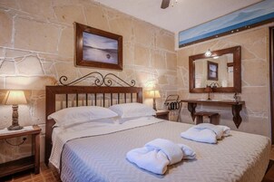 Un dormitorio con pared de piedra, una cama con ropa blanca, una mesita de noche con una lámpara, un cabecero de madera y un espejo sobre el tocador.