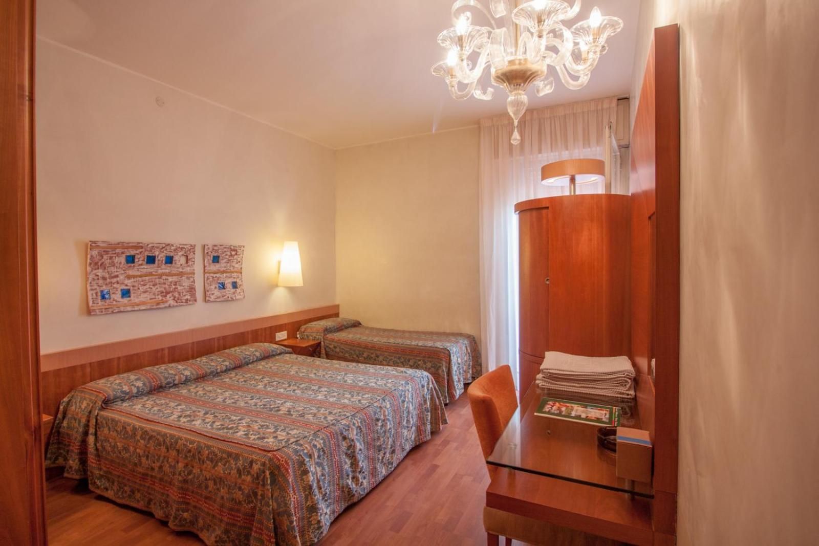 A hotel room with two beds, a wooden wardrobe, a glass table, and a chandelier.