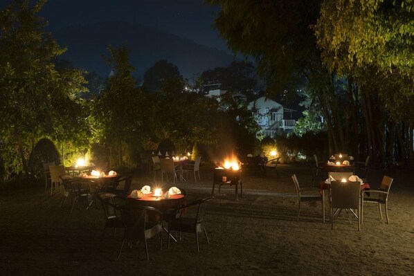 Un área de comedor al aire libre con mesas y sillas dispuestas para los huéspedes, rodeada de árboles y un edificio distante.