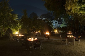 Un espace de restauration en plein air, aménagé avec des tables et des chaises pour les clients, entouré d’arbres et avec un bâtiment visible au loin.