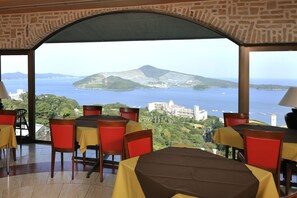 A restaurant with tables and chairs, a view of a body of water and a mountain, and a stone wall. - Altia Toba (Toba)