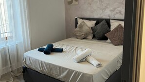 A neatly made bed with pillows and a rolled towel, a wall with a decorative piece, and a window with sheer curtains. - Elegant and cozy; apparently recently renovated (Béziers)