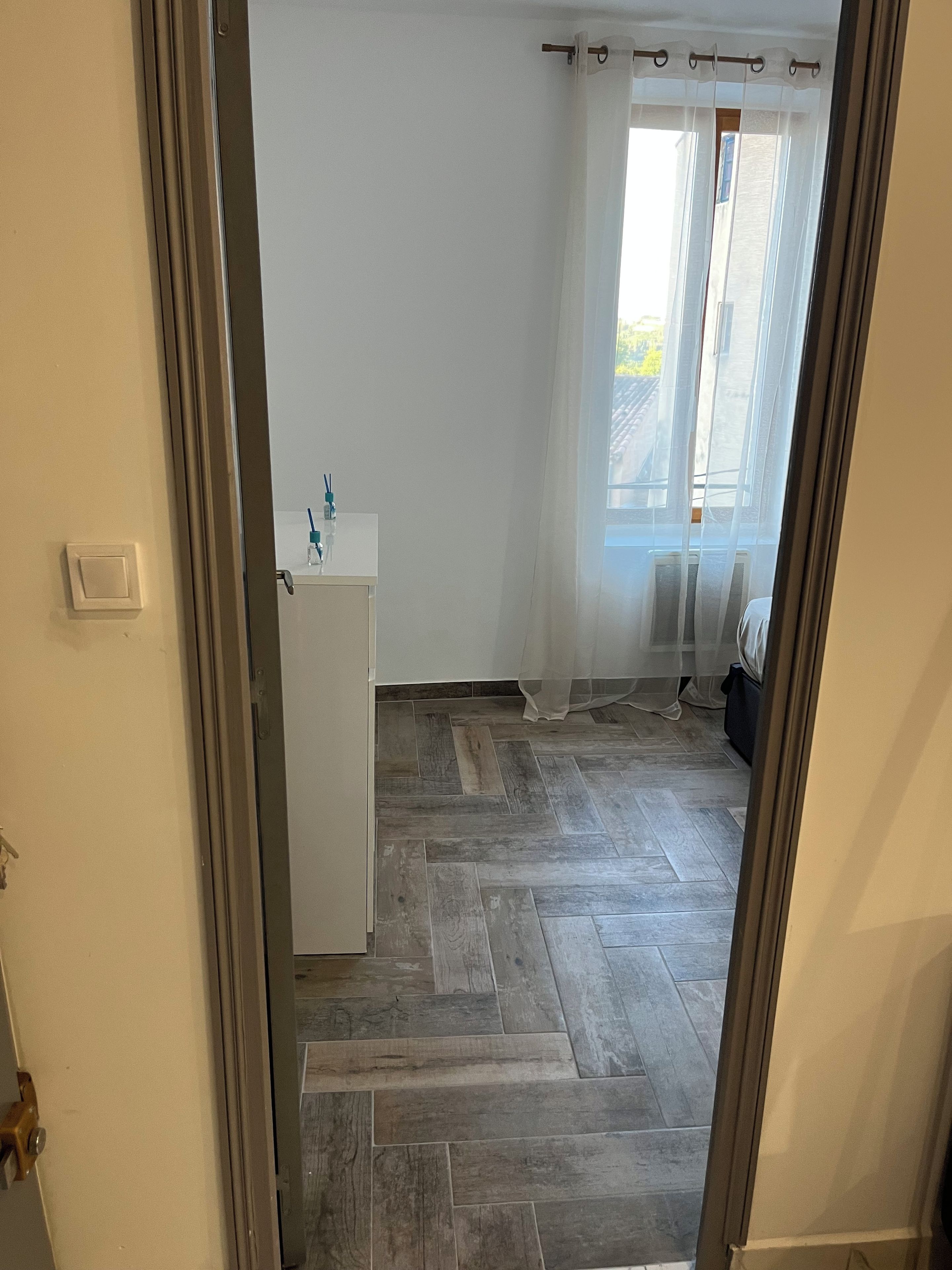 Une pièce dotée d’un parquet à motifs en chevrons, d’une armoire blanche et d’une fenêtre ornée de rideaux légers.