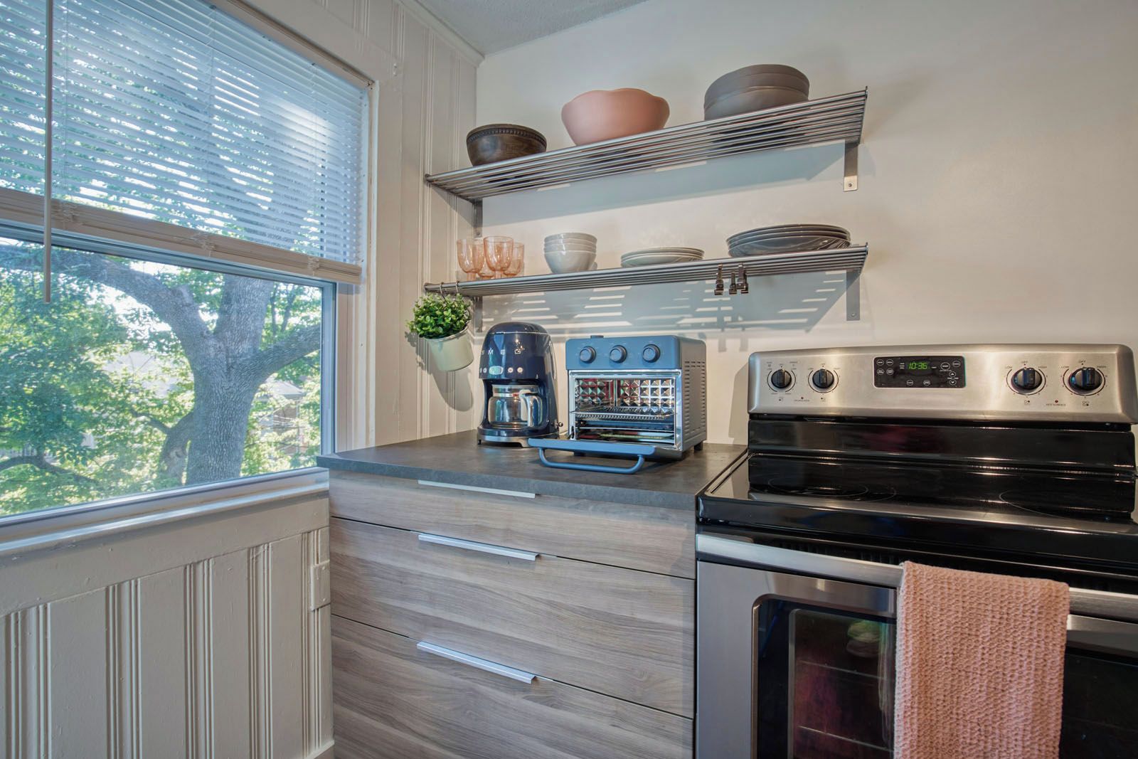 A kitchen with a built-in oven, a coffee maker, and a toaster. There are shelves above the counter holding dishes and a hanging plant.