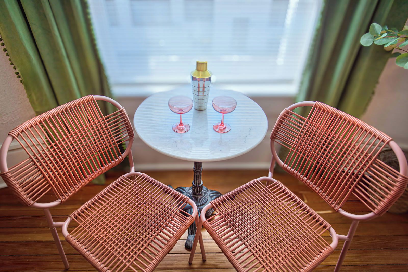 A round table with two pink wicker chairs, two wine glasses, and a bottle of water.