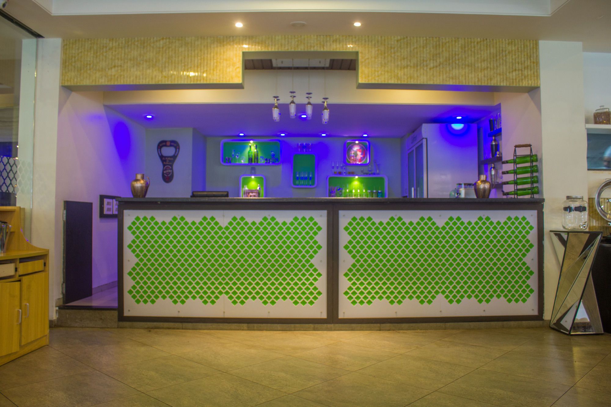 Un espace bar avec un mur vert à motifs, un comptoir où sont exposées différentes bouteilles et un plafond doté d’un éclairage encastré.