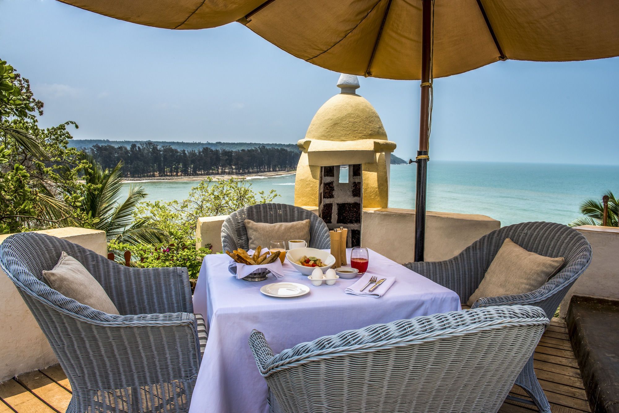 A dining area with wicker chairs and a table set for two, overlooking a beach and ocean.