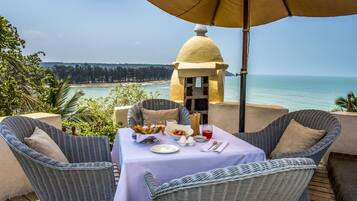 A dining area with wicker chairs and a table set for two, overlooking a beach and ocean.