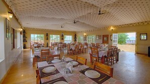 Un comedor espacioso con varias mesas dispuestas para comidas, ventiladores de techo y grandes ventanales que dejan entrar luz natural.