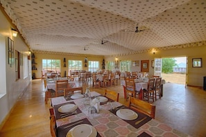 A spacious dining area with multiple tables set for meals, ceiling fans, and large windows allowing natural light. - Wind Desert Camp (Jaisalmer)