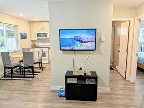 A modern living room with a flat-screen TV mounted on the wall, a dining area with a table and chairs, and a kitchen in the background.