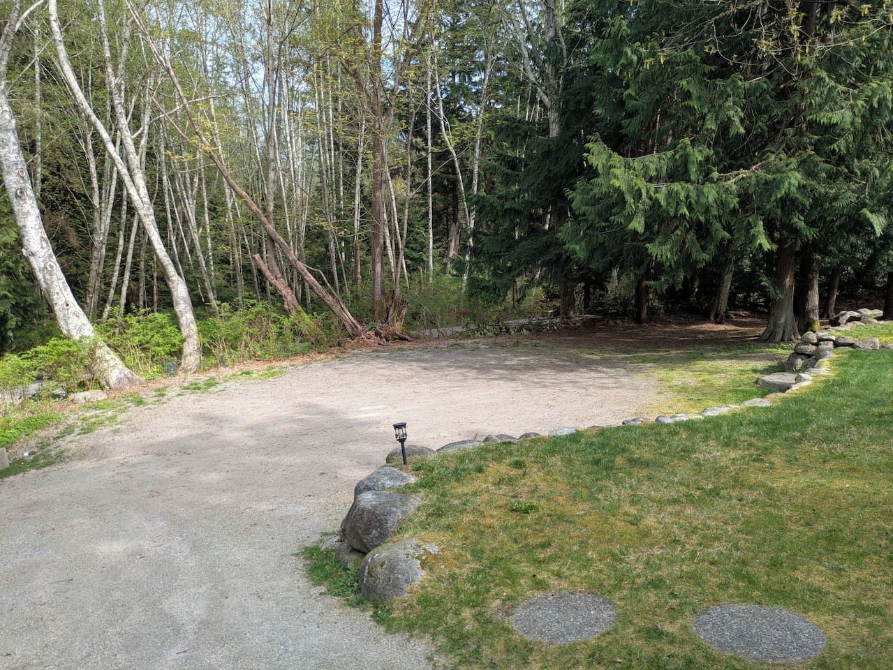 Un chemin de gravier mène à une zone abritée, où se trouve un lampadaire, le tout étant entouré d’arbres et de rochers.