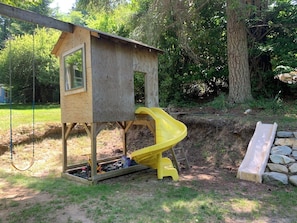 Una casita de juegos de madera con un tobogán amarillo, un columpio y una escalera.