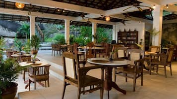 A spacious indoor dining area with wooden tables and chairs, surrounded by potted plants and a view of the outdoors.