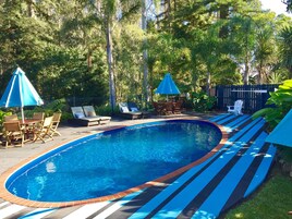 Una piscina con borde de ladrillo y cubierta a rayas azules, rodeada de sillas de descanso y sombrillas.