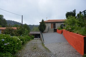 Ein Kopfsteinpflasterweg führt zu einem Steinhaus mit Ziegeldach, umgeben von Grünflächen und einer roten Mauer.