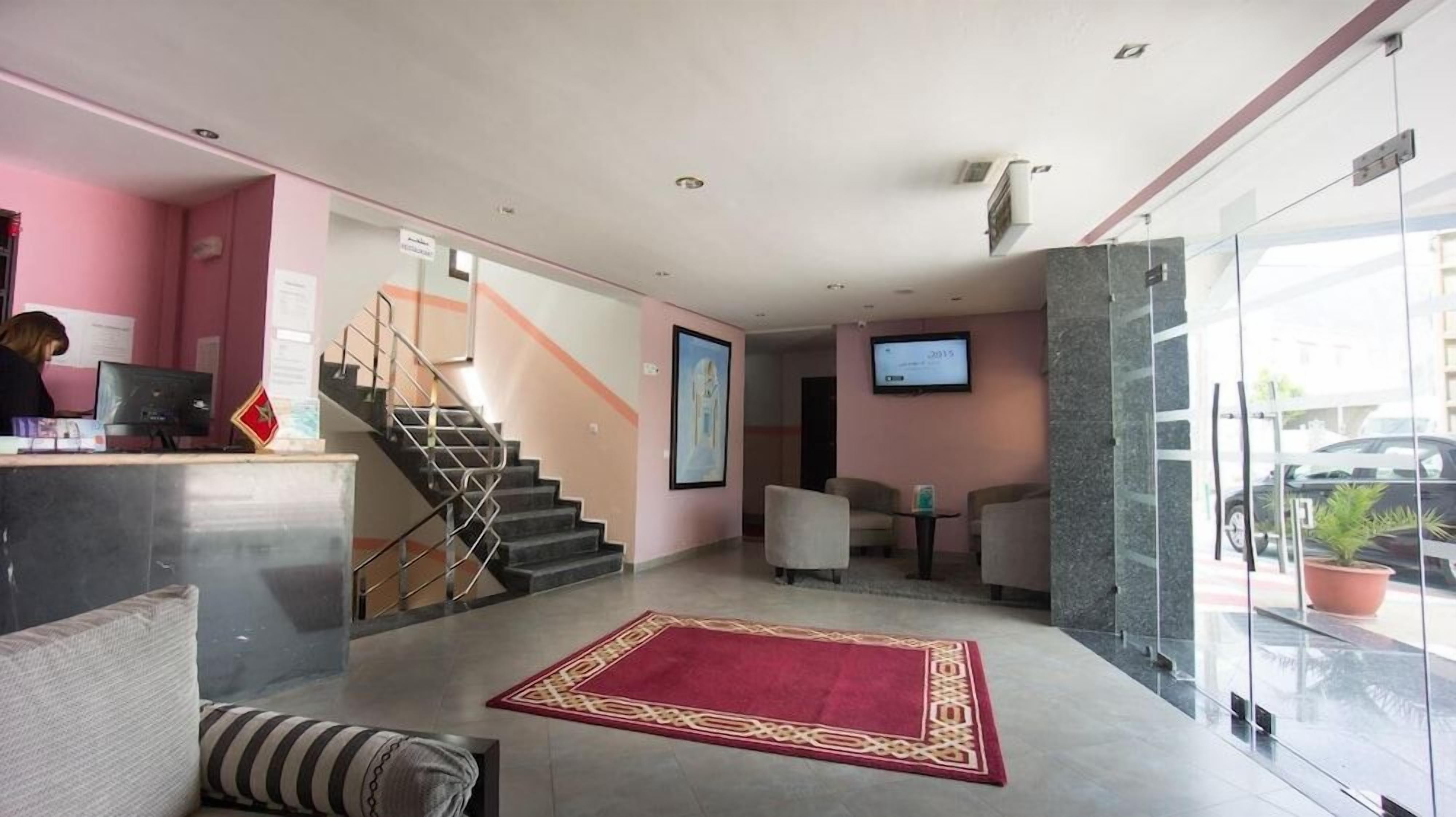 Un vestíbulo de hotel con mostrador de recepción, una escalera, zona de estar y una pantalla de televisión.