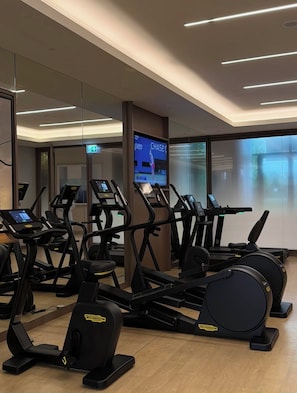 A gym with multiple cardio machines, a digital display, and a glass wall.