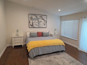 A bedroom with a bed, nightstands, a rug, a painting, and a window with blinds.