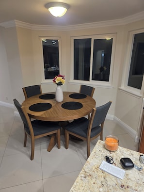 A dining area with a round wooden table, six chairs, a vase with flowers, and a candle on a marble countertop.