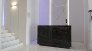 A modern reception desk with a marble top, a staircase with white steps, and a wall-mounted light fixture. - Akrogiali Boutique Hotel (Kassandra)