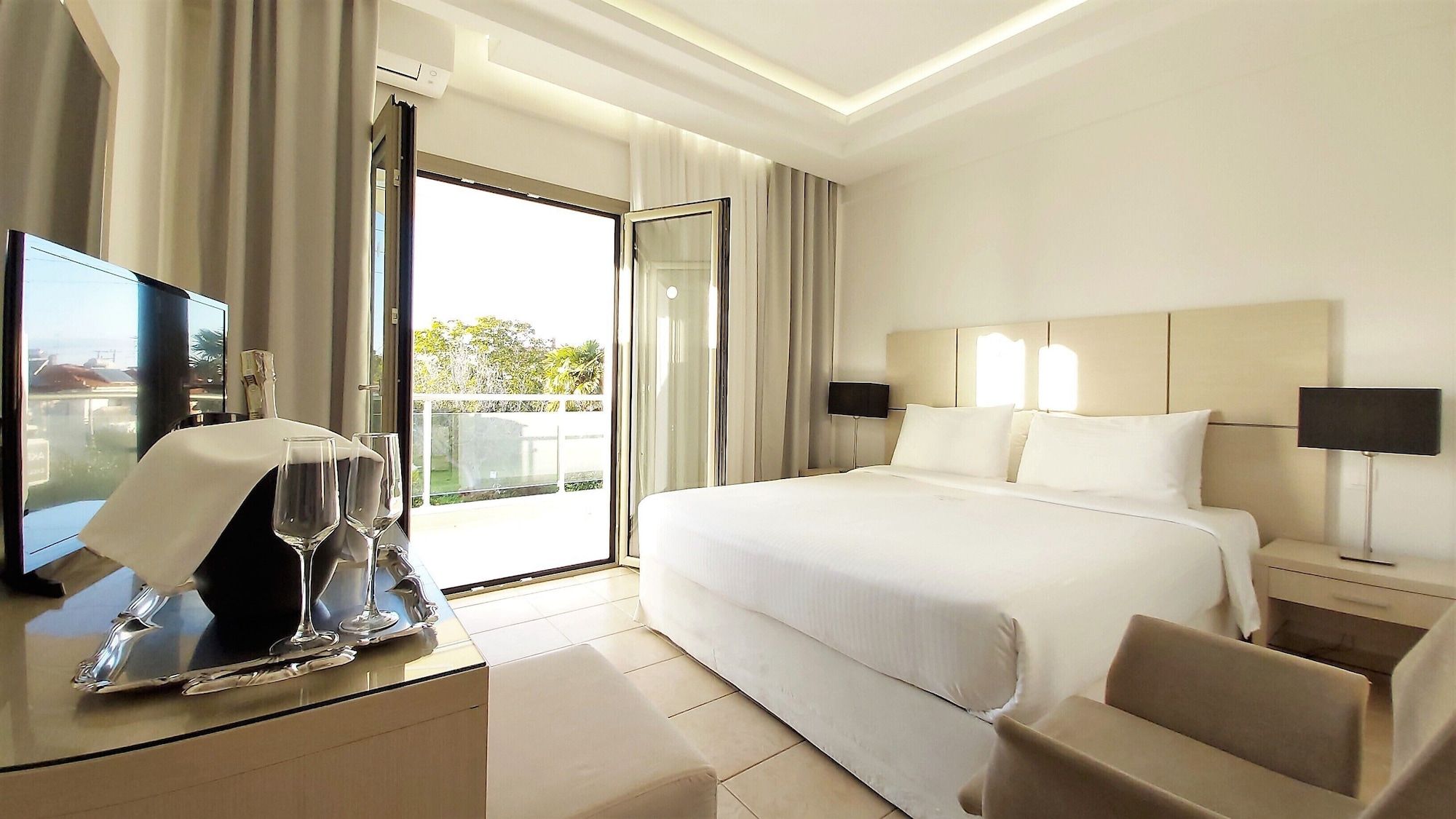 A hotel room with a large bed, a TV, a desk with a glass of water, and a view of the outdoors.