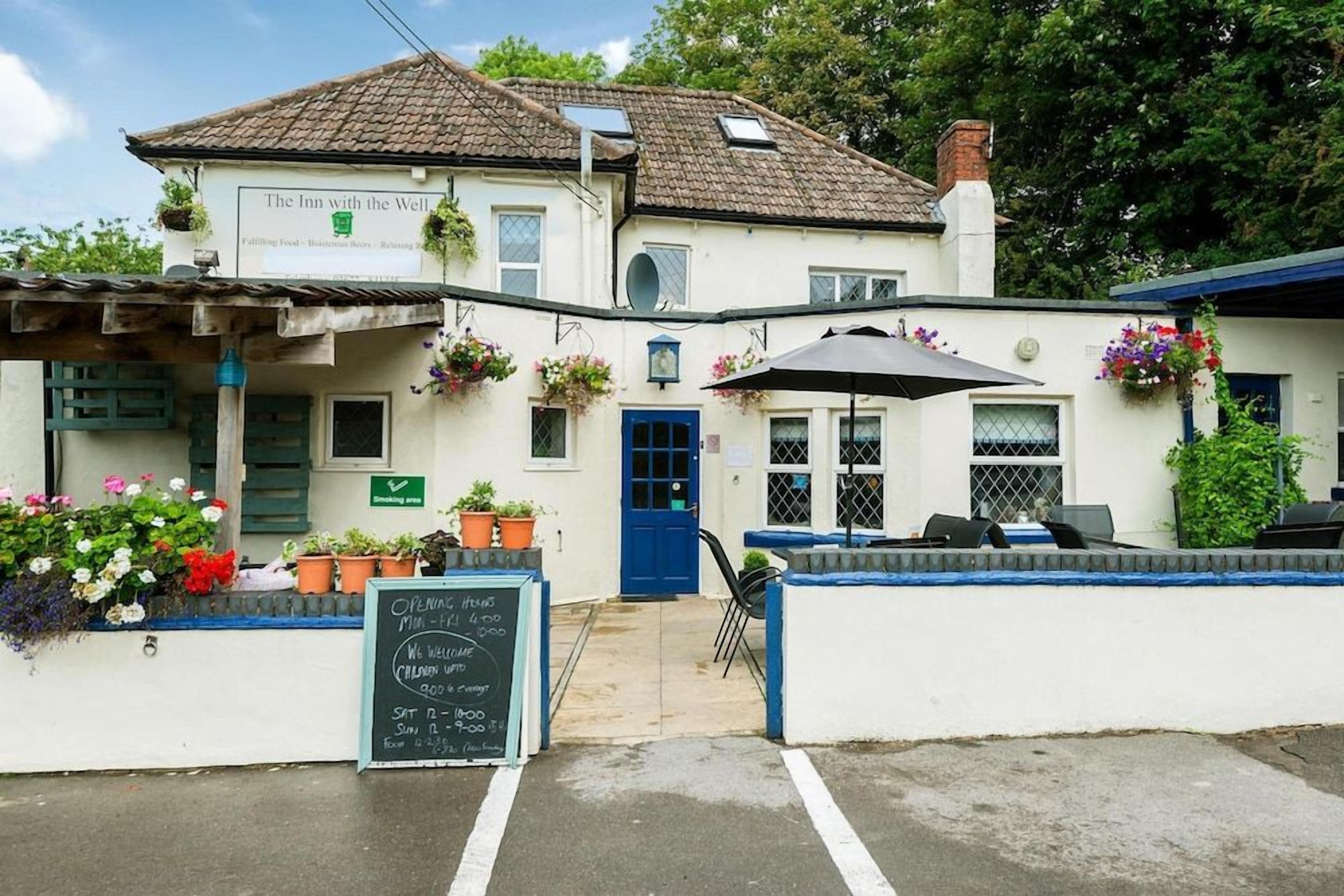 A white building with a blue door, outdoor seating, and a signboard with opening hours.