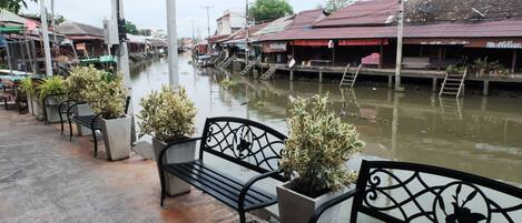 A riverside walkway with benches and potted plants, overlooking a waterway with buildings on the opposite bank.