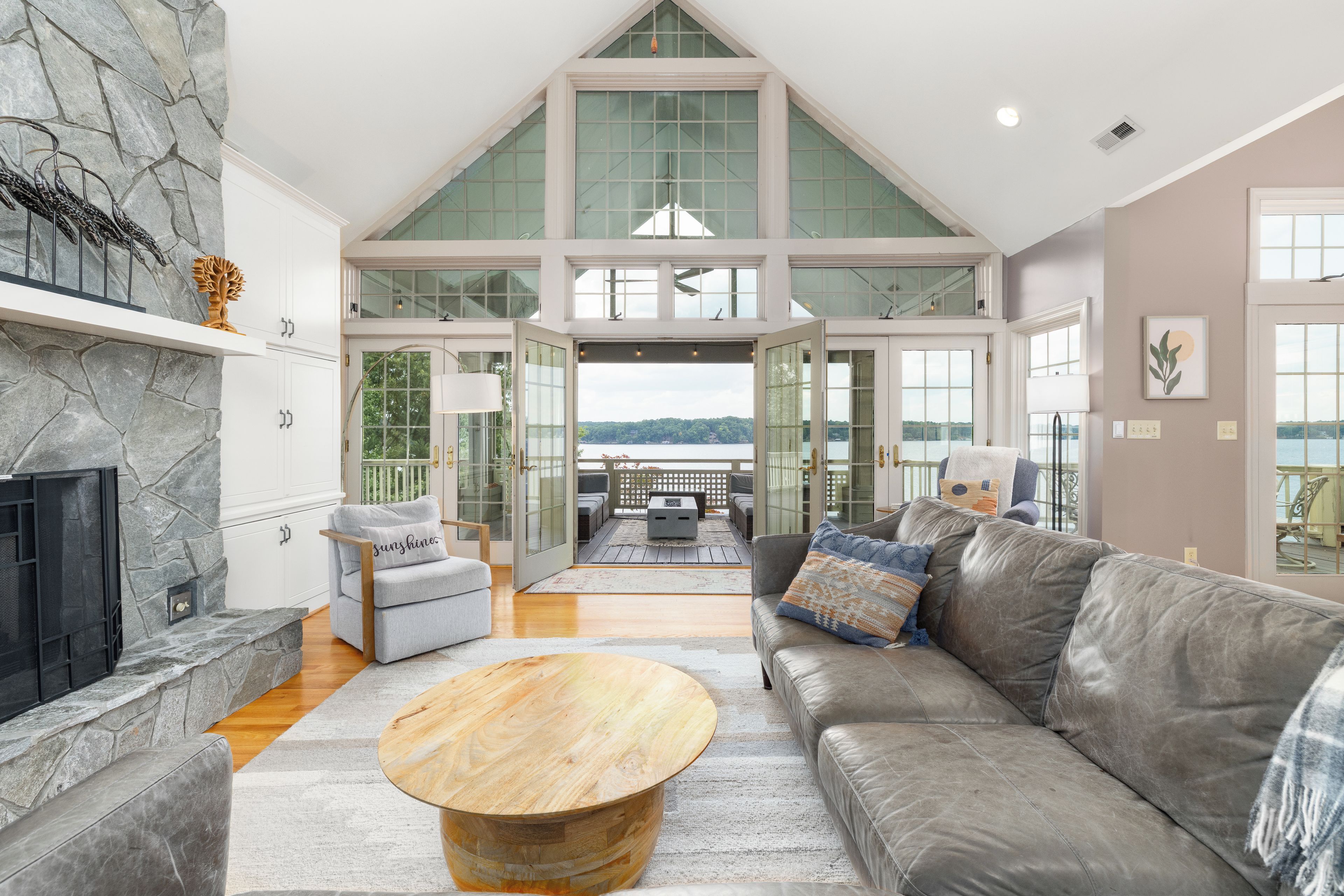 A spacious living area with a stone fireplace, a grey sofa, a wooden coffee table, and a view of the outdoors through large windows.