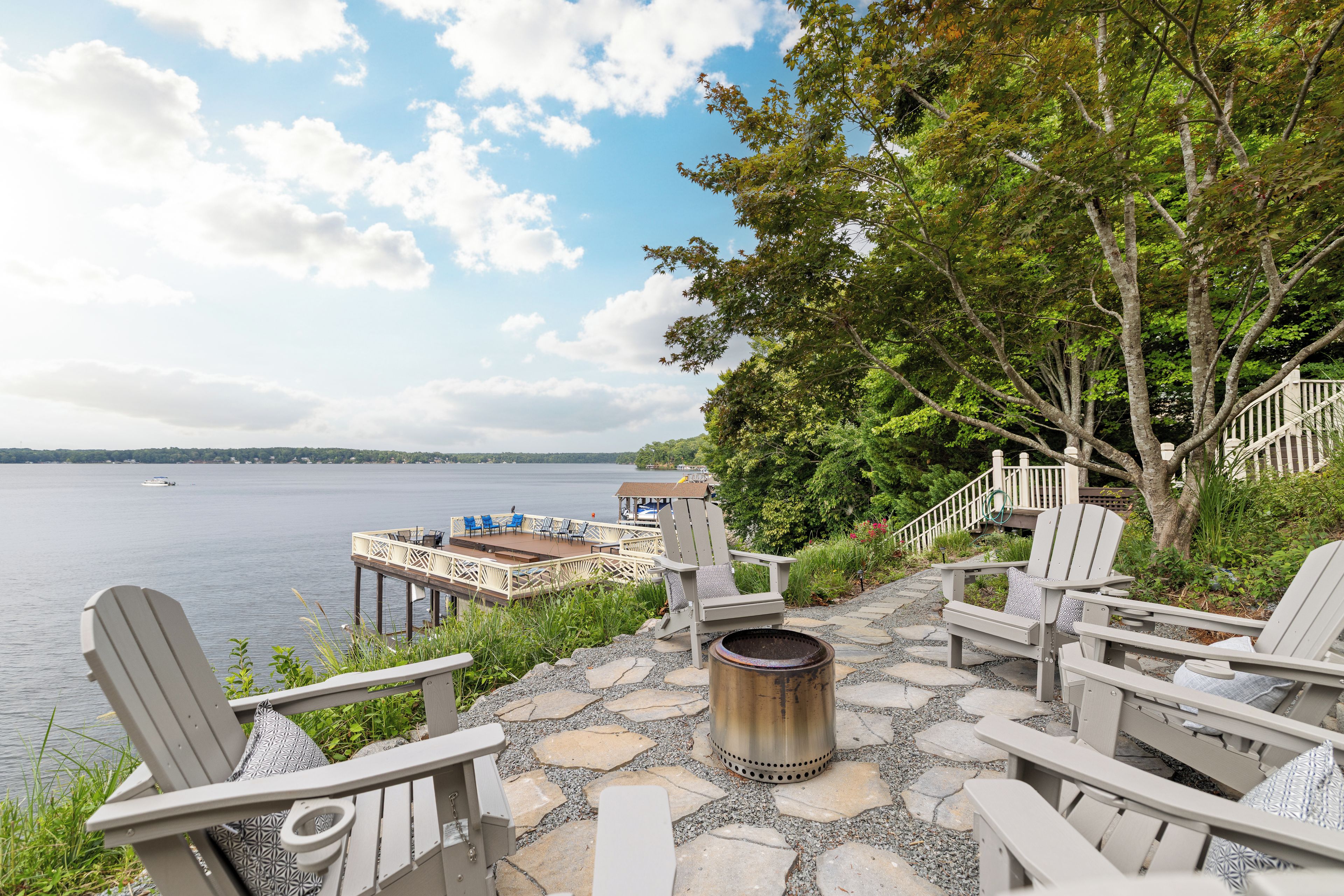 A deck with Adirondack chairs and a fire pit overlooking a lake with a dock and boat in the distance.
