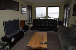 A living room with a large flat-screen TV, a wooden coffee table, a black leather sofa, and a view of a body of water through large windows. - Studioat10 (Hobart)