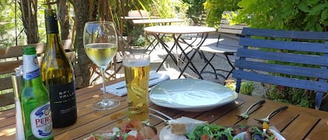 Una mesa de madera al aire libre con una botella de vino, una copa de vino blanco, un plato y diferentes alimentos como queso, pan y una ensalada.