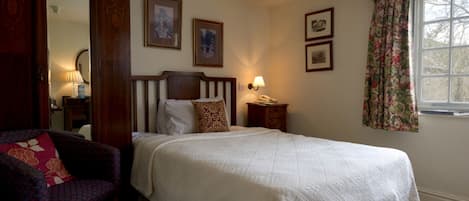 A bedroom with a large bed, a chair, a bedside table with a lamp, and a window with curtains.
