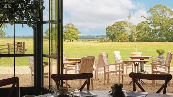 Un comedor con sillas y mesas de madera, vista a un campo de césped y una valla de madera.