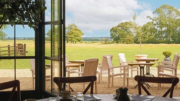 Un comedor con sillas y mesas de madera, vista a un campo de césped y una valla de madera.