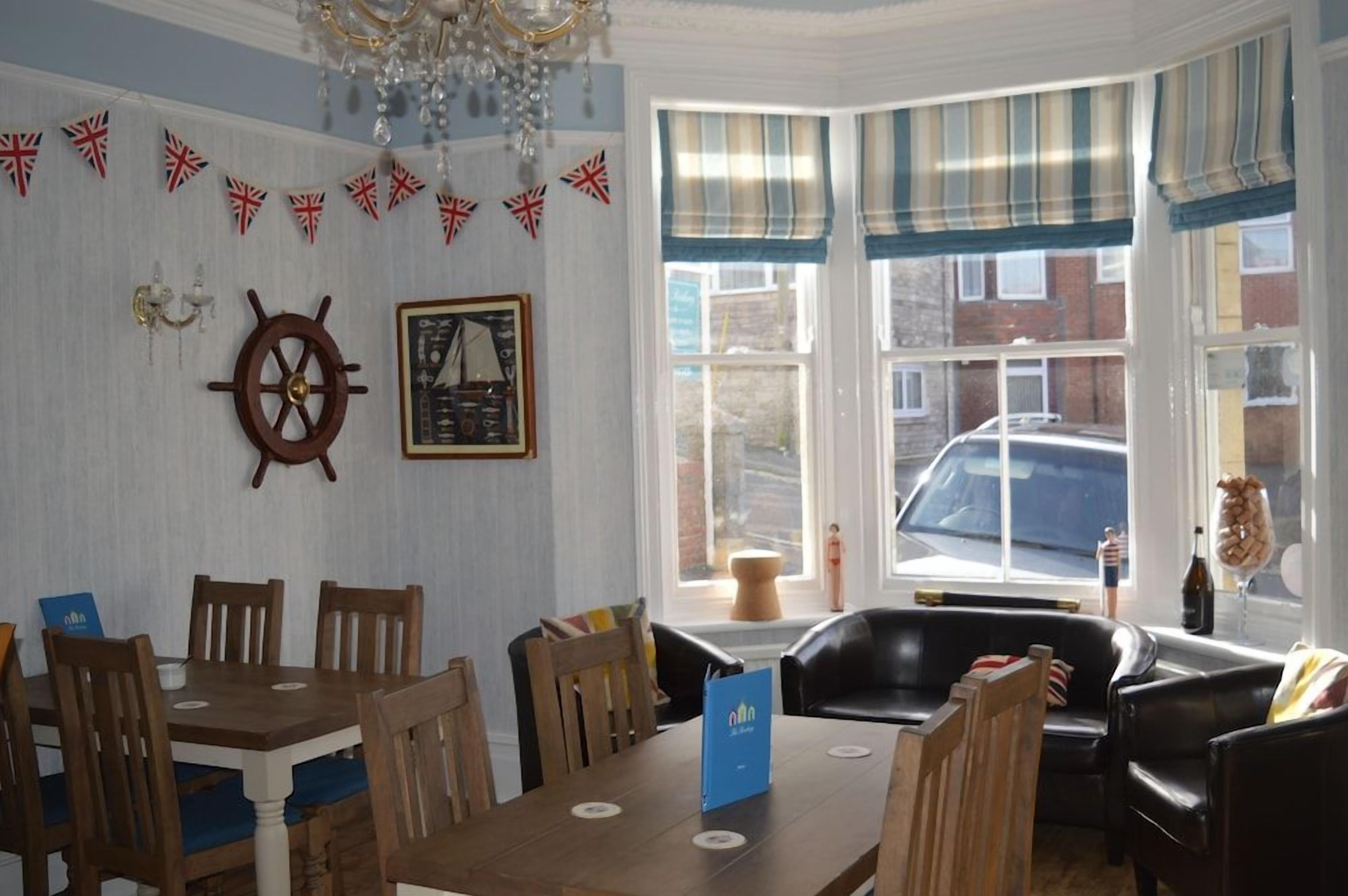 A room with a dining table, chairs, a black leather sofa, a ship's wheel decoration, and a window with a view of the outside.