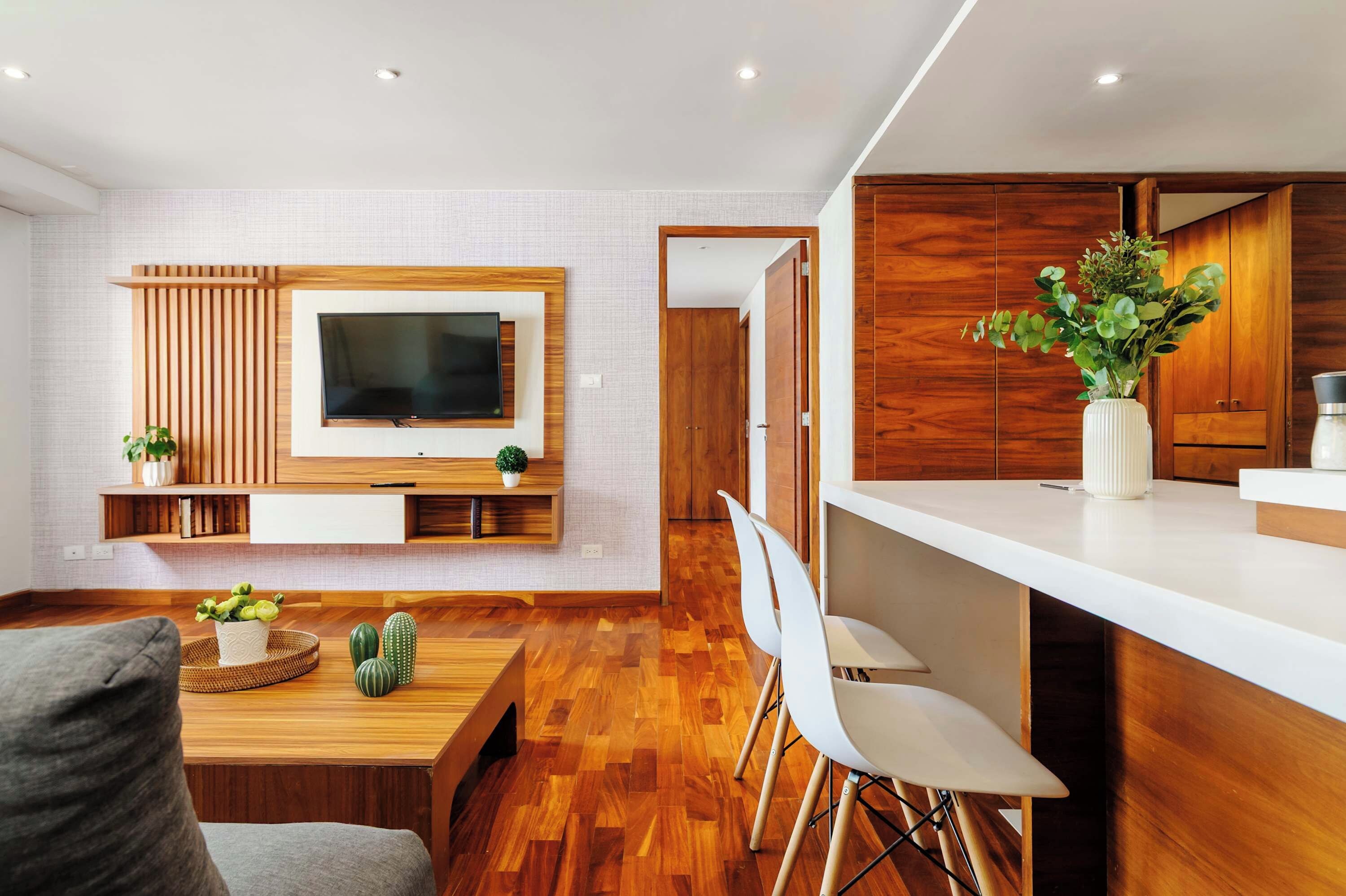 Un salon moderne avec un téléviseur à écran plat fixé à un mur en bois, un coin repas avec une table et des chaises en bois, et une cuisine avec un comptoir blanc et des armoires en bois.