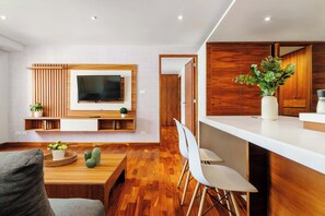 A modern living room with a flat-screen TV mounted on a wooden wall, a dining area with a wooden table and chairs, and a kitchen area with a white countertop and wooden cabinets. - w* | Luxury & Cozy 1BR in Prime Polanco (Ciudad de México)