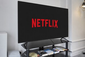A flat-screen TV mounted on a black stand with the word 'NETFLIX' in red letters. Below the TV, there is a black shelf with books and a white trash bin. - Family & Pet Friendly Flat | Free Parking&Wi-fi (Ceredigion)