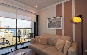 Ein Wohnzimmer mit einer Couch, einem Tisch, einem Stuhl, einer Stehlampe und einem großen Fenster mit Blick auf Gebäude.