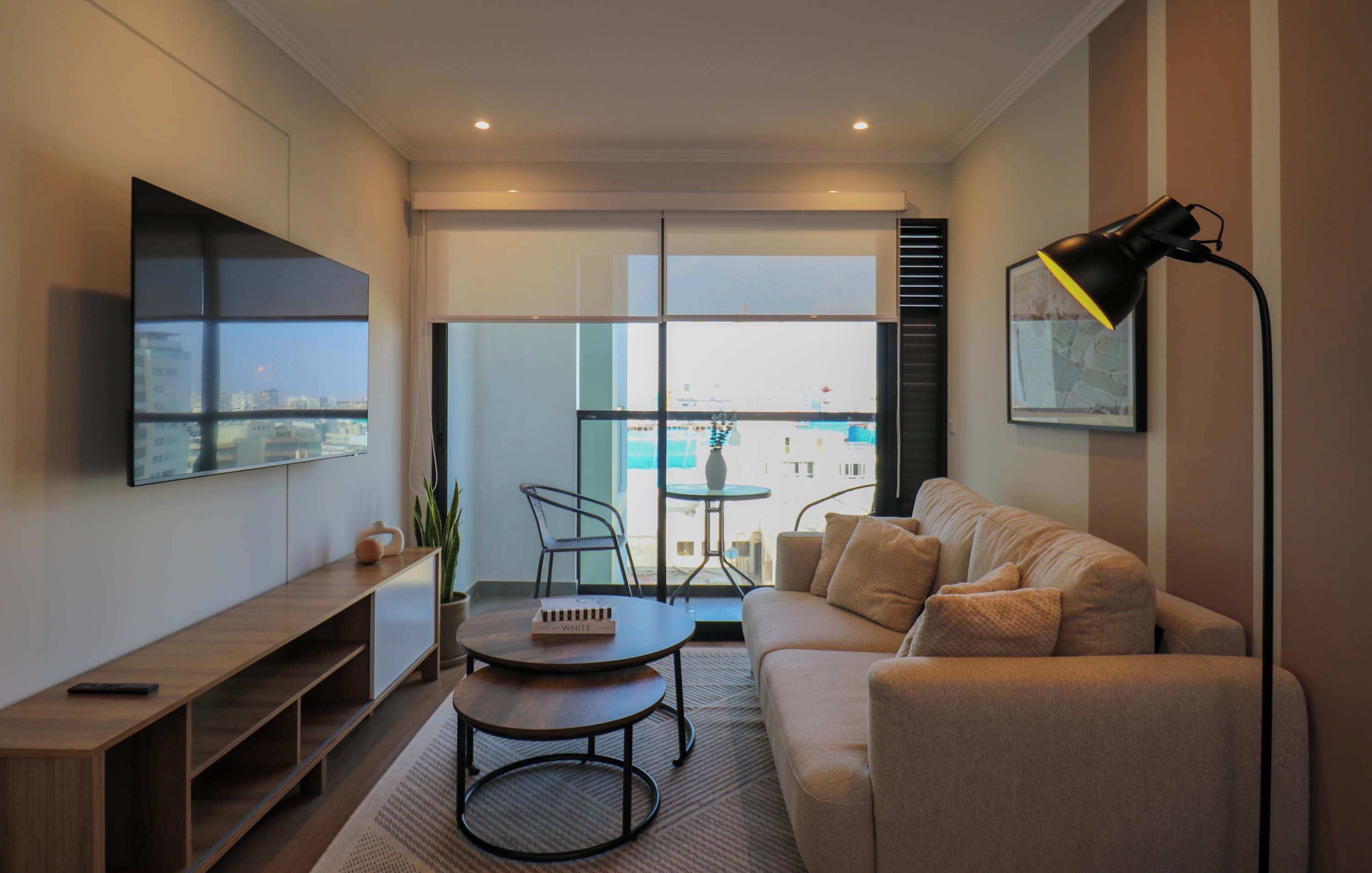 Un salon moderne avec un canapé, une table basse, un téléviseur fixé au mur et un balcon offrant une vue sur la ville.