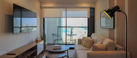 A modern living room with a sofa, a coffee table, a TV mounted on the wall, and a balcony with a view of the city.