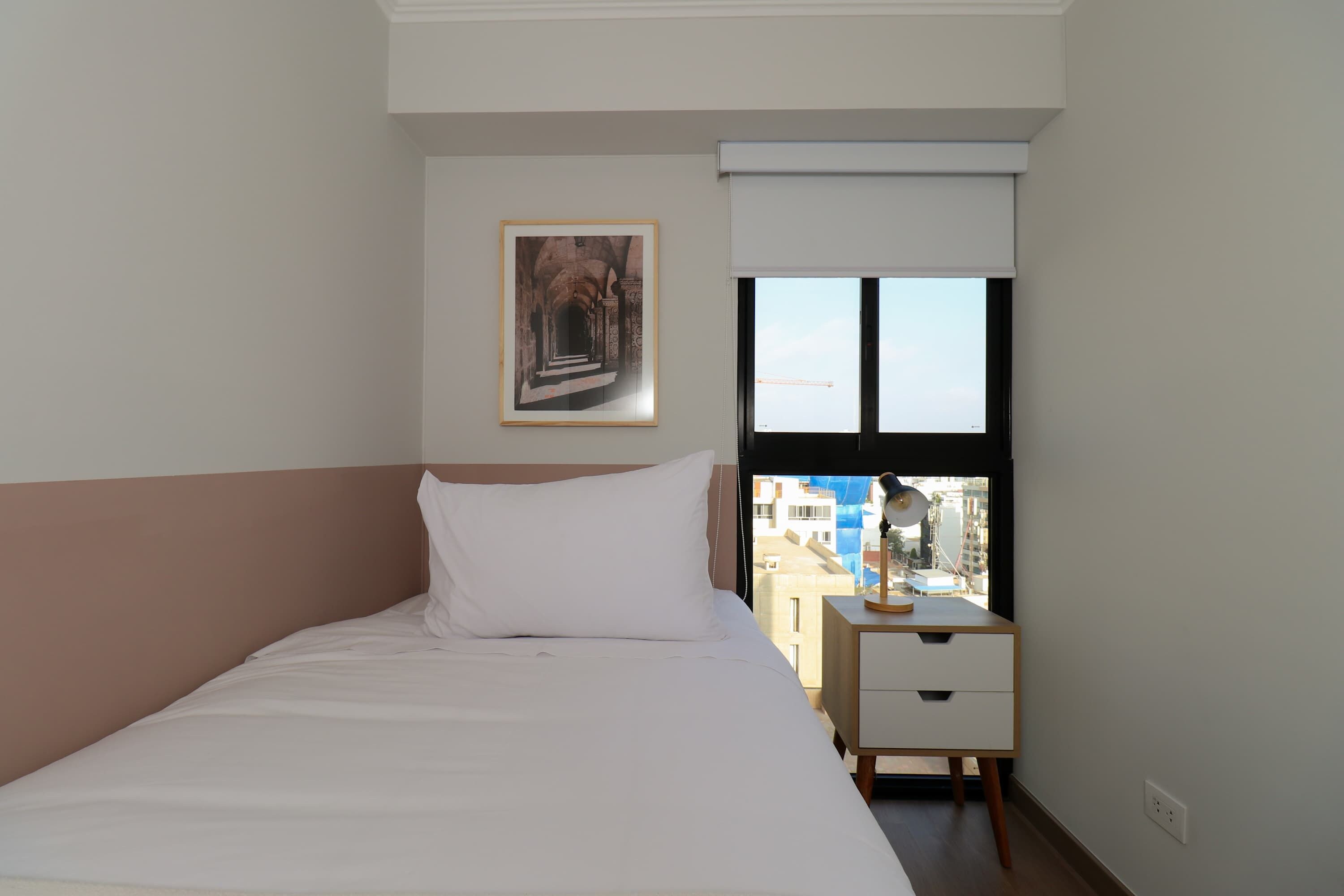 Une chambre à coucher avec un lit, une table de chevet, une fenêtre donnant sur des immeubles et un tableau encadré accroché au mur.
