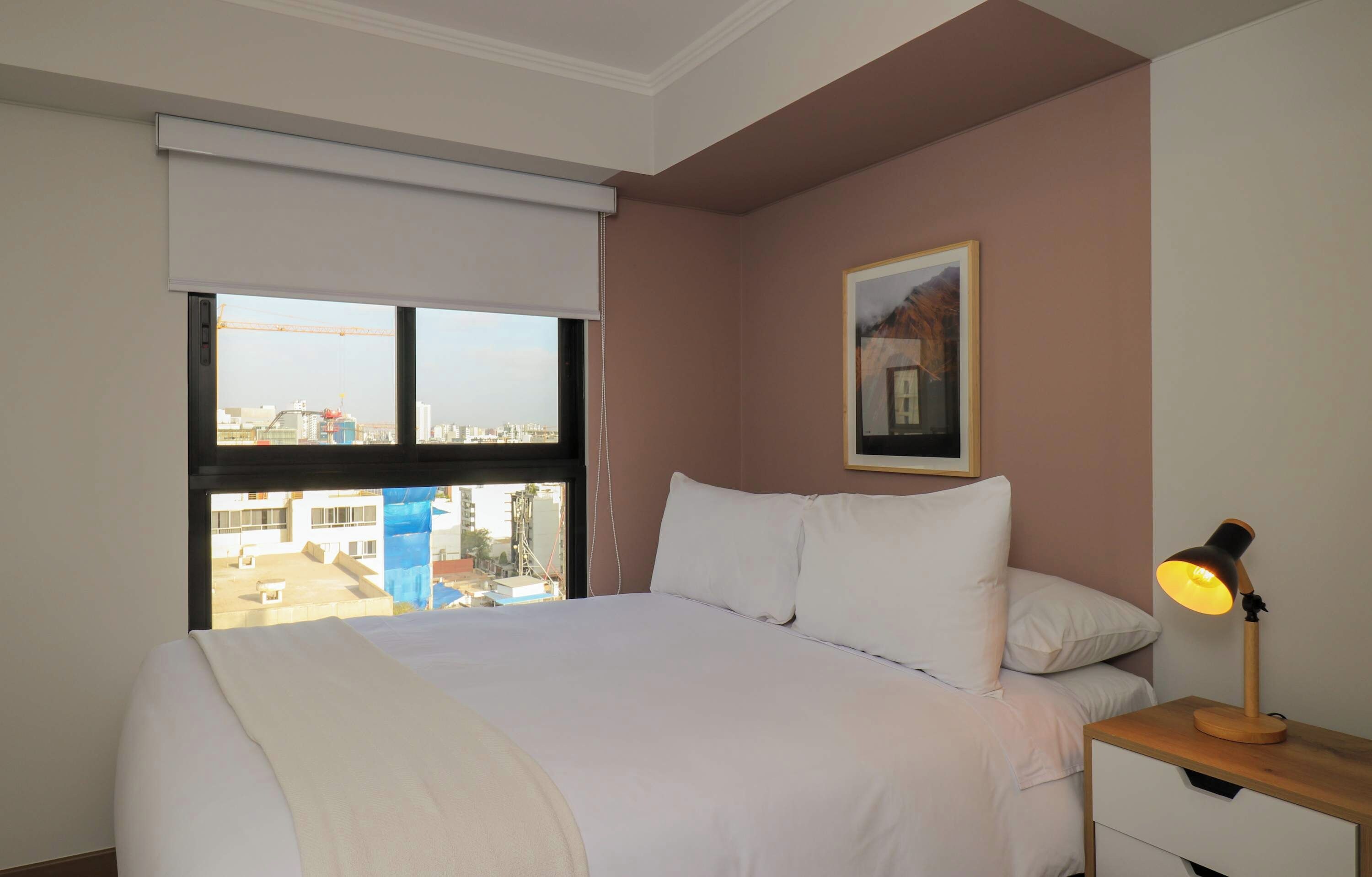 A hotel room with a bed, a nightstand with a lamp, a window with a view of buildings, and a framed picture on the wall.