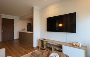 Ein modernes Wohnzimmer mit Flachbildfernseher an der Wand, Holzboden, einem Esstisch mit einer Zeitschrift und einer Küchenzeile inklusive Kühlschrank und Schränken.