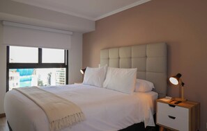 A hotel room with a large bed, two bedside lamps, a nightstand with a lamp, and a view of buildings through the window. - w* | Deluxe 3BR w/ Balcony in Prime Miraflores (Miraflores)