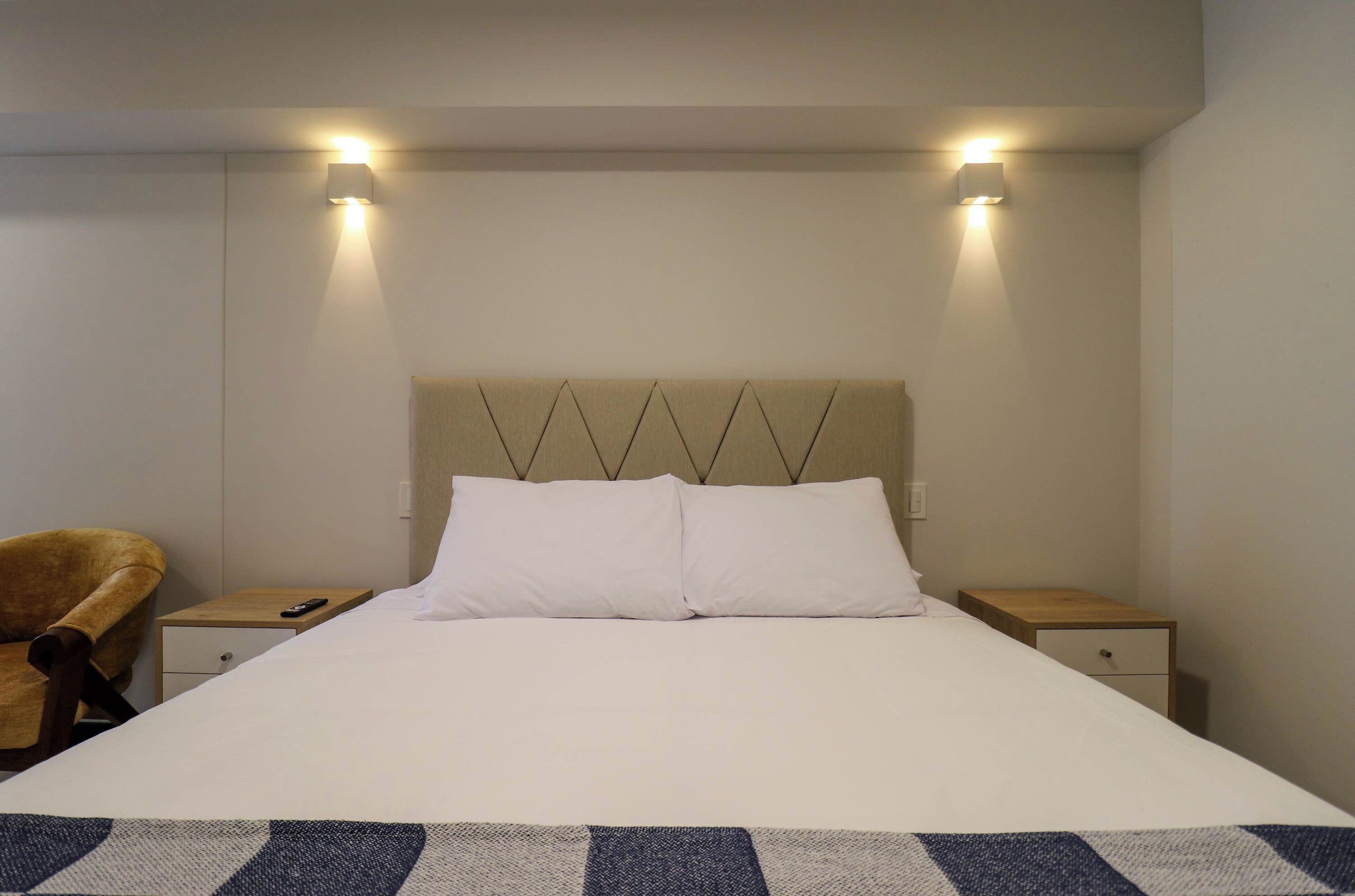 Una habitación de hotel con una cama, dos mesitas de noche, una silla y dos luces fijadas a la pared.
