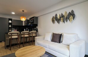 A modern living room with a sofa, a coffee table, and a dining area with chairs and a kitchen in the background. - w* | Modern 2BR w/ Balcony in Prime Miraflores (Miraflores)