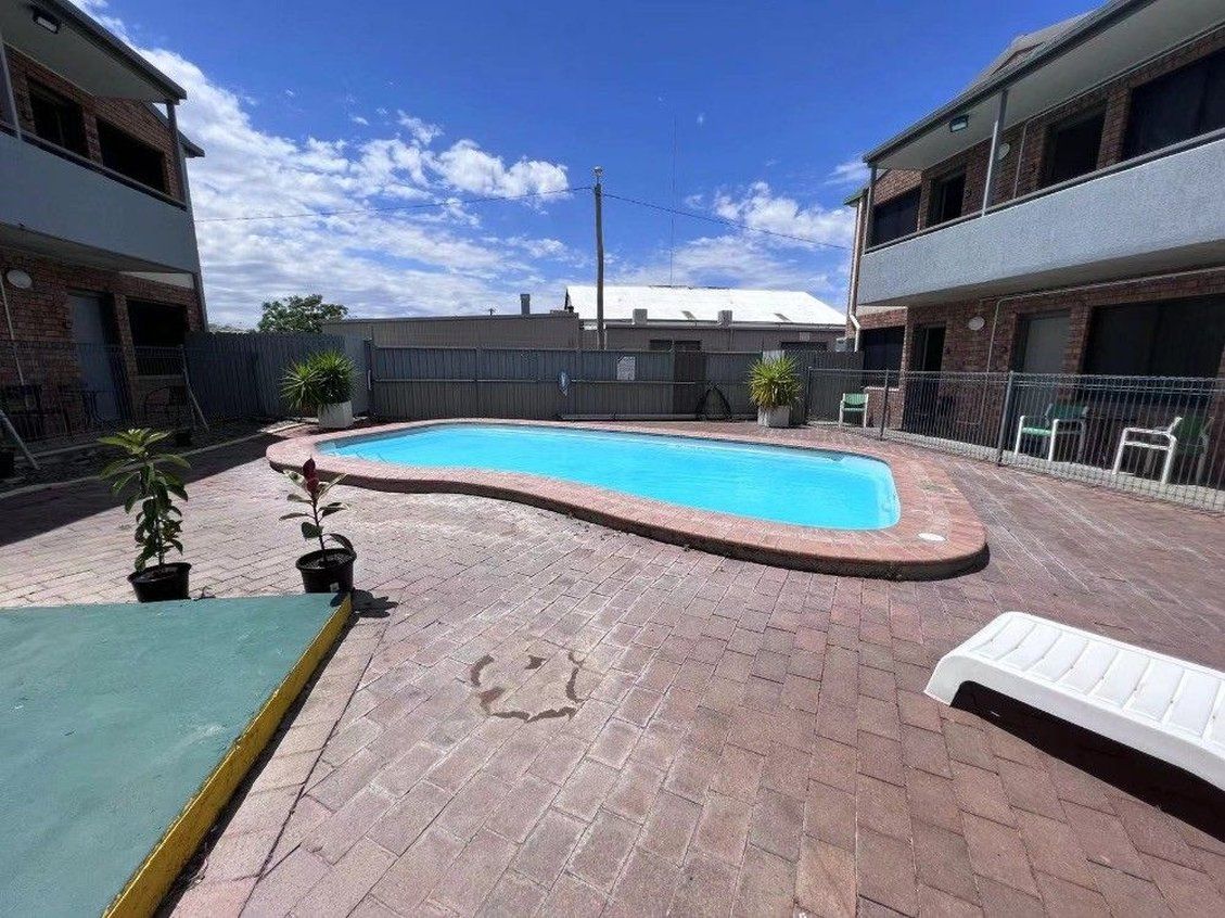 A rectangular swimming pool with a brick border, surrounded by a paved patio with potted plants.